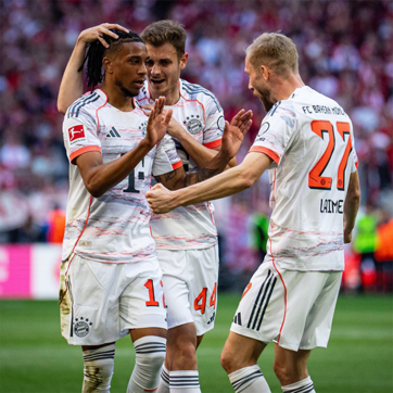 Three football players in Bayern Munich jerseys celebrate a goal on the pitch, showing excitement and camaraderie. One player with dreadlocks receives a pat on the head, while another gestures enthusiastically. The background features a cheering crowd, adding to the lively atmosphere of the moment.