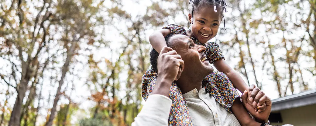 A joyful man smiles up at a young girl on his shoulders, both wearing matching floral outfits. The girl is laughing happily, capturing a moment of playful connection in a natural outdoor setting with blurred trees in the background.
