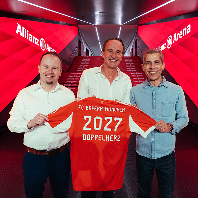 Three men are standing together inside Allianz Arena, holding a red FC Bayern München jersey. The jersey features "2027" and "Doppelherz" printed on the back. The background shows illuminated walls and stairs, emphasizing a celebratory atmosphere.