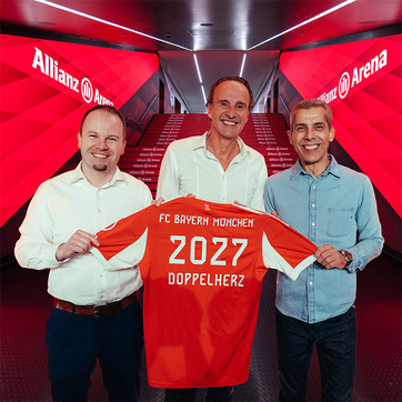 Three men are standing together inside Allianz Arena, holding a red FC Bayern München jersey. The jersey features "2027" and "Doppelherz" printed on the back. The background shows illuminated walls and stairs, emphasizing a celebratory atmosphere.