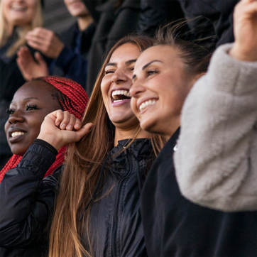 Three young women are smiling and laughing together. They show enthusiasm and joy, possibly cheering for an event. The woman on the left has short black hair and is wearing a black jacket, while the others have long hair and are clad in casual outfits.