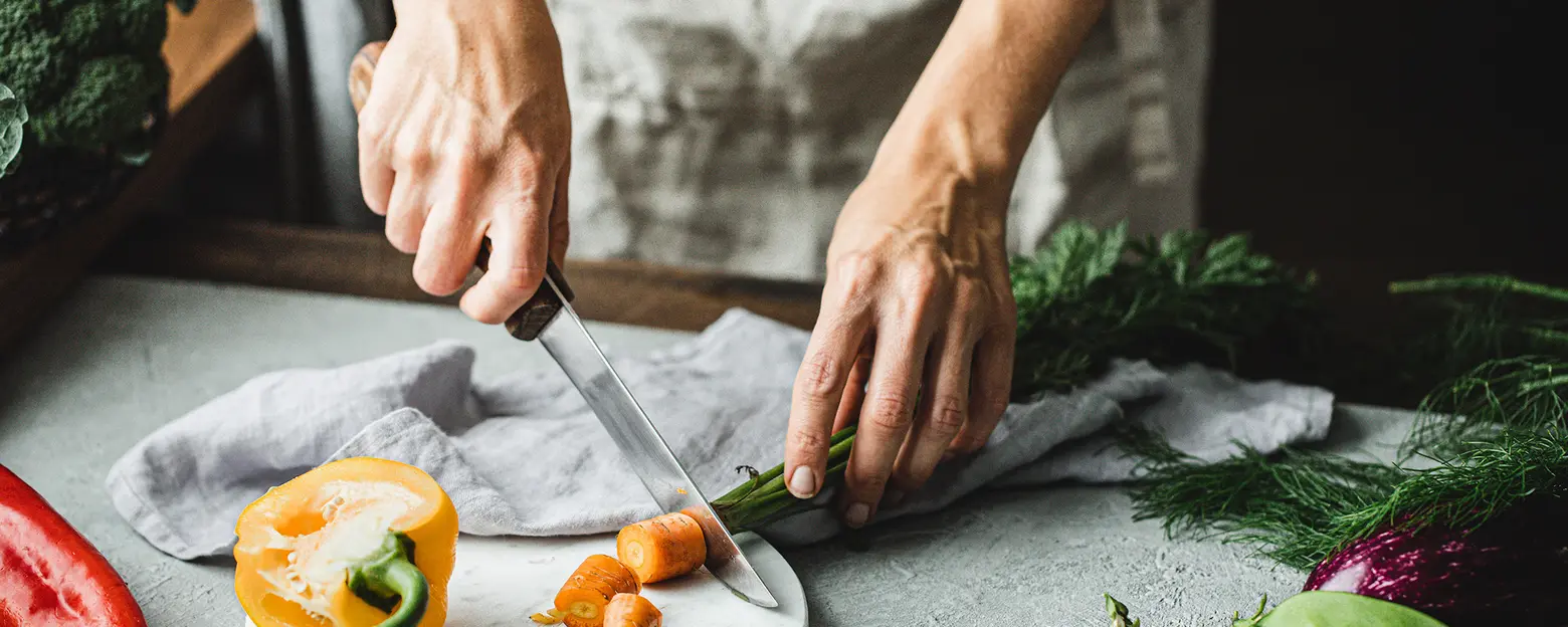 A person's hands are seen slicing orange carrots atop a light-colored cutting board. Nearby, a yellow bell pepper and green herbs are also visible, creating a vibrant kitchen scene focused on food preparation.