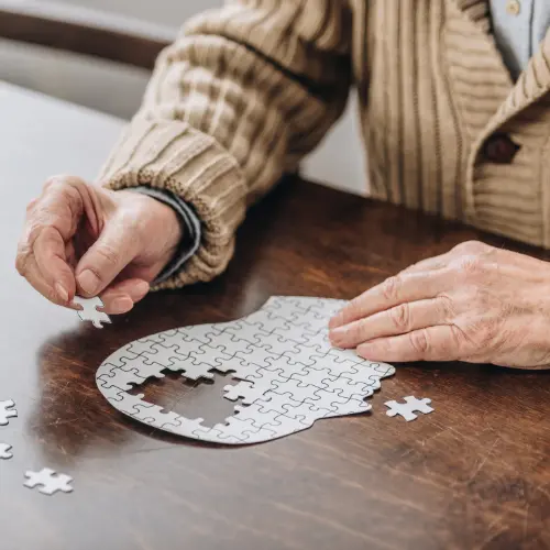 An elderly person is assembling a white jigsaw puzzle on a wooden table. They are focused on placing a puzzle piece into the nearly complete silhouette shape, with several scattered pieces around. The scene conveys concentration and engagement in the activity.