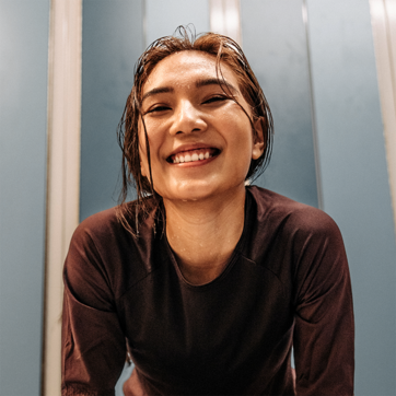 A smiling woman with wet hair leans forward, exuding a sense of joy and energy. She is wearing a dark athletic top, suggesting an active lifestyle. The background features light blue walls, complementing her cheerful expression.