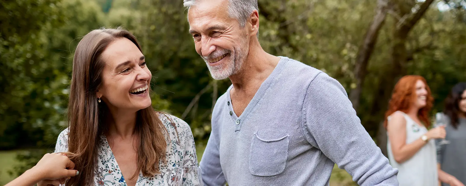 A man and a woman laugh together outdoors, enjoying each other's company. The woman has long brown hair and wears a floral blouse, while the man has gray hair and a light gray sweater. They share a moment of joy amidst greenery.